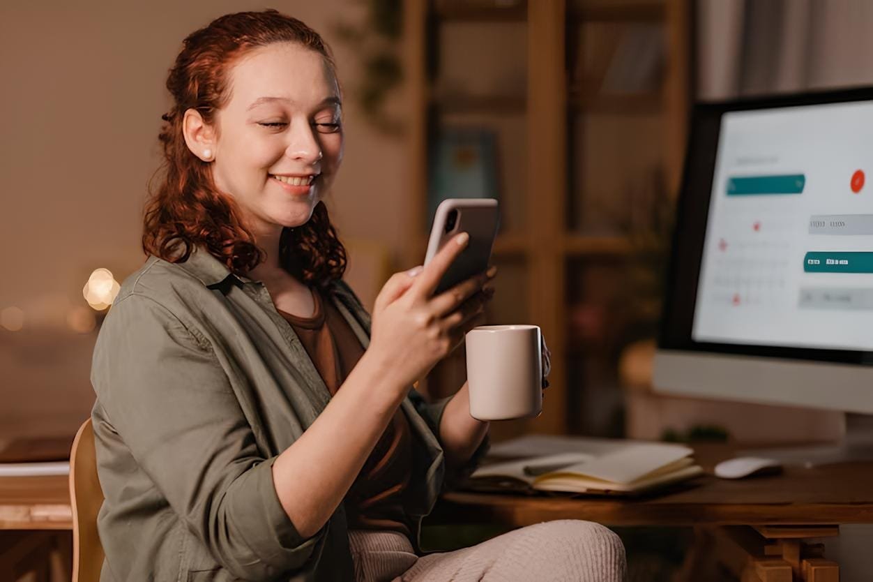 mulher em casa usando smartphone na frente do computador enquanto toma cafe 23 2148793444 1