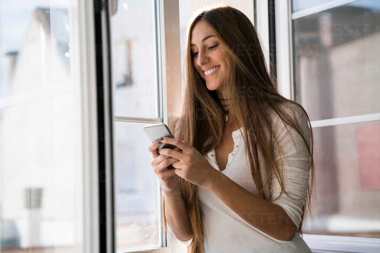 smiling young woman looking at cell phone at the window KKAF00325